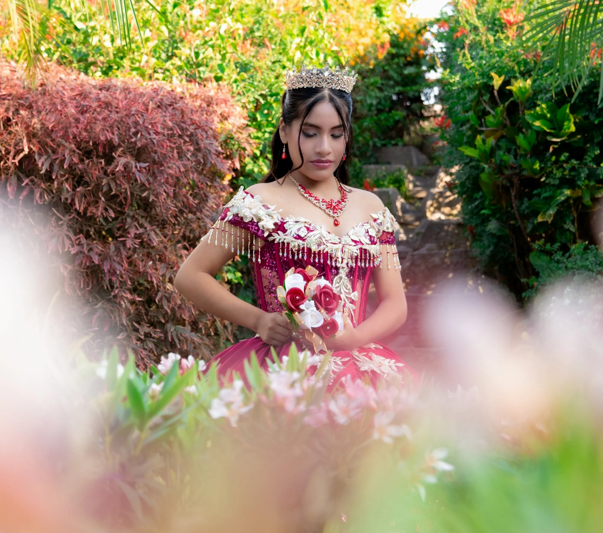 Quinceañera con corona, vestido fucsia y ramo de flores en jardín